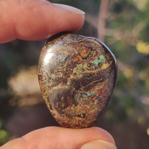 May include: A polished, irregular, rounded stone featuring a dark brown base color with intricate, swirling patterns of metallic copper, deep red, and vibrant turquoise blue inclusions throughout its smooth, reflective surface.