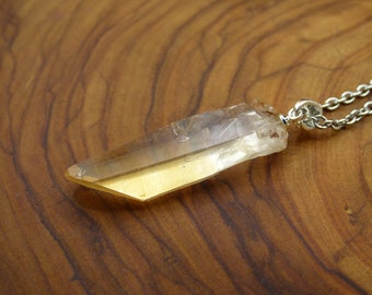 May include: A natural, raw crystal pendant necklace. The crystal is a light amber color with a pointed shape, suspended from a silver-colored chain. The necklace is displayed on a wooden surface.