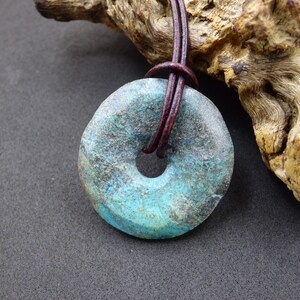 Puede incluir: Un colgante de piedra azul turquesa con un cordón de cuero marrón. La piedra es redonda con un agujero en el centro y tiene una superficie rugosa y texturizada.