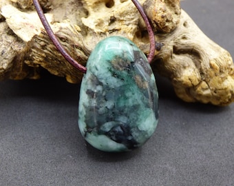 May include: A polished, teardrop-shaped green emerald pendant with dark inclusions and a smooth, glossy surface. The gemstone is suspended from a thin, dark brown leather cord that loops through the top of the stone, creating a simple necklace.