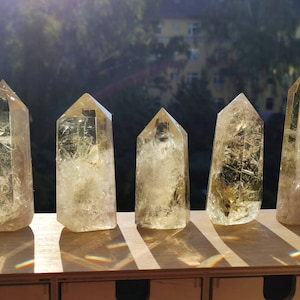 Könnte beinhalten: Fünf klare Quarz-Kristallspitzen unterschiedlicher Größe, angeordnet auf einer Holzoberfläche. Die Kristalle haben einen hellgelben Farbton und innere Einschlüsse, die das Sonnenlicht einfangen. Der Hintergrund zeigt eine verschwommene Ansicht von Bäumen und Gebäuden.