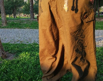 May include: Olive green loose-fitting trousers featuring a drop-crotch design, side cargo pockets with button closures, and dark floral screen-printed patterns on the front. The pants have a relaxed silhouette and a textured fabric appearance.