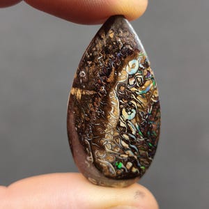 Könnte beinhalten: Ein tropfenförmiger Boulder-Opal-Edelstein mit einer dunkelbraunen Eisensteinmatrix, der komplizierte, wirbelnde Muster aus irisierenden grünen, blauen und goldenen Blitzen auf der gesamten polierten Oberfläche aufweist.