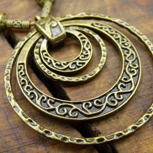 Könnte beinhalten: Eine goldfarbene Halskette mit einem Anhänger, der drei konzentrische Kreise mit aufwendigen Designs zeigt. Die Kreise sind durch ein kleineres, verziertes Stück verbunden.