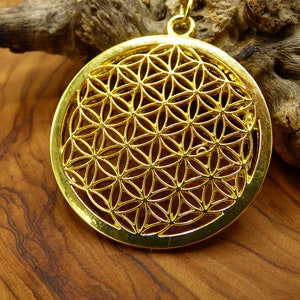 May include: A gold-colored pendant necklace featuring a round, intricate design known as the Flower of Life. The pendant is hanging on a chain against a wooden background.