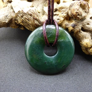 Pu&ograve; includere: Un ciondolo in giada verde scuro a forma di ciambella, appeso a un cordoncino in pelle marrone.