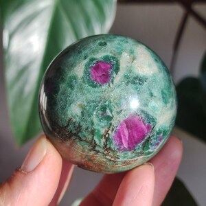 Puede incluir: Una esfera pulida de zoisita verde con vibrantes inclusiones de rubí rosa. La gema se sostiene en una mano, mostrando su superficie lisa y reflectante. Los colores de la esfera varían del verde oscuro a tonos más claros, con el rubí agregando un contraste llamativo.