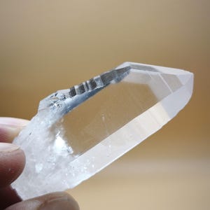 May include: A clear, transparent quartz crystal point with a sharp, faceted termination. The crystal features natural horizontal striations along one side and a textured, opaque base, showcasing internal clarity and light refraction.