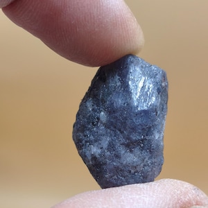 May include: A raw, unpolished sapphire gemstone with a deep blue-gray color and a textured, irregular surface. The stone features natural facets and crystalline structures throughout its body, displaying a matte finish with subtle light reflections.