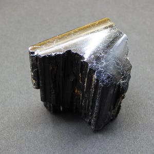 Può includere: Un cristallo di tormalina nera grezza con una superficie superiore lucida e riflettente. Il cristallo ha un aspetto ruvido e strutturato con striature visibili e una forma naturale. Il bordo superiore ha una lucentezza metallica.