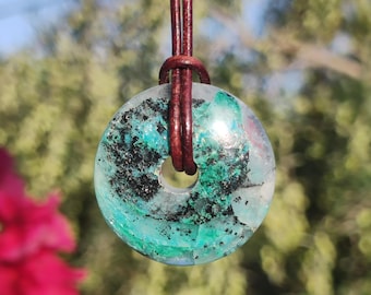 May include: A circular pendant featuring a polished stone with vibrant green and black mineral inclusions, suspended from a dark brown leather cord. The stone has a smooth, translucent surface with a central hole through which the cord passes.
