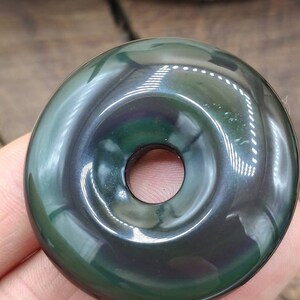 K&ouml;nnte beinhalten: Ein dunkler gr&uuml;ner Obsidian-Donut-Anh&auml;nger mit einem Loch in der Mitte. Der Anh&auml;nger hat eine gl&auml;nzende, irisierende Oberfl&auml;che.