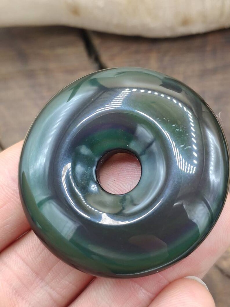 K&ouml;nnte beinhalten: Ein dunkler gr&uuml;ner Obsidian-Donut-Anh&auml;nger mit einem Loch in der Mitte. Der Anh&auml;nger hat eine gl&auml;nzende, irisierende Oberfl&auml;che.