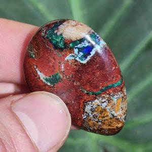 May include: A polished, oval-shaped gemstone with a rich, earthy color palette. The stone features a deep reddish-brown base, accented with streaks of vibrant green, patches of white, and hints of blue and yellow. The stone is held by a finger.
