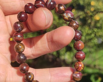 May include: A beaded bracelet featuring round, polished beads in shades of brown, red, and gold. The bracelet is strung together, creating a flexible band. The beads have a marbled appearance, with variations in color and pattern.