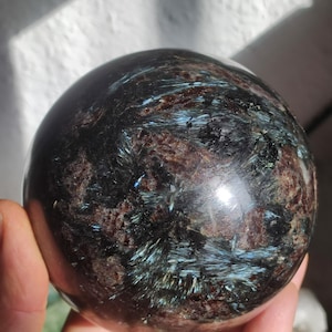 Op de afbeelding: Een gepolijste zwarte bol met iriserende blauwe en groene strepen. De bol is gemaakt van een natuurlijke steen, mogelijk een soort aventurijn.