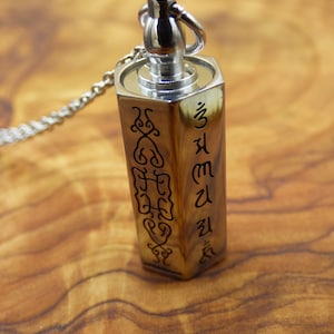 Könnte beinhalten: Eine silberfarbene Halskette mit einem rechteckigen Anhänger in Holzoptik. Der Anhänger ist mit schwarzen Symbolen und Text verziert. Der Anhänger hängt an einer silbernen Kette. Der Hintergrund ist eine Holzoberfläche.