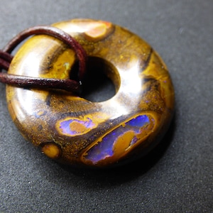 May include: A brown and gold opal pendant with a purple and blue iridescent sheen. The pendant is in the shape of a donut and is strung on a brown leather cord.