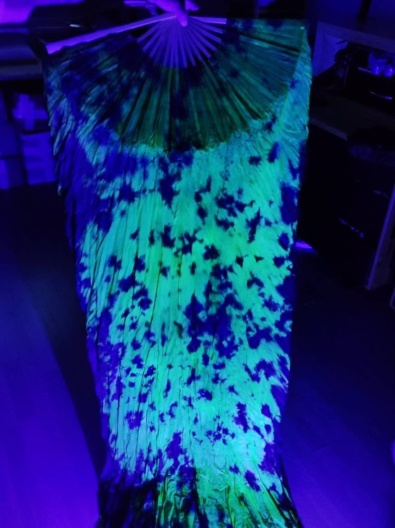 May include: A vibrant, open hand fan with a tie-dye pattern. The fan features a neon green and blue color scheme, with dark blue splotches across the surface. The fan's wooden ribs are visible at the top.