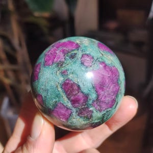 Può includere: Una sfera lucida di rubino zoisite, una gemma dall'aspetto screziato. La sfera è principalmente verde acqua con ampie macchie irregolari di magenta intenso. La pietra è tenuta in mano, con lo sfondo sfocato.