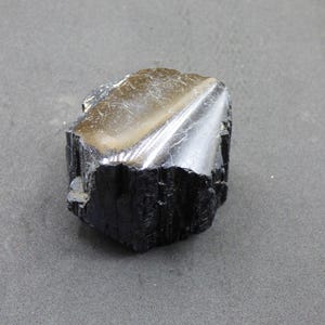 Può includere: Un cristallo grezzo di tormalina nera naturale con una superficie lucida e riflettente. Il cristallo ha una base scura e strutturata e una parte superiore lucida e metallica. La pietra è posta su uno sfondo grigio.