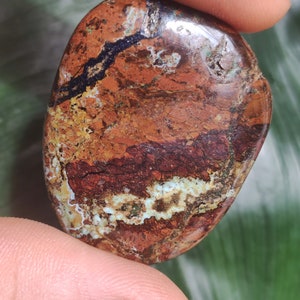 May include: A polished, rounded stone featuring a complex pattern of deep red, earthy brown, and ochre tones, accented by thin, jagged veins of dark navy blue and bright white mineral deposits that create a textured, multi-layered appearance.