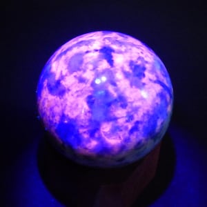 Può includere: Un oggetto sferico luminoso con un aspetto marmorizzato, che mostra una miscela di tonalità viola e blu. L'orbita sembra essere illuminata dall'interno, creando un effetto visivo accattivante. L'oggetto è posto su uno sfondo scuro.