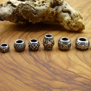 Puede incluir: Ocho cuentas de metal plateado con diseños intrincados. Las cuentas están dispuestas en una fila sobre una superficie de madera.
