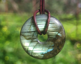 May include: A round, polished labradorite pendant with a central hole, strung on a brown leather cord. The stone displays iridescent blue, green, and gray hues. The pendant is set against a blurred green background.