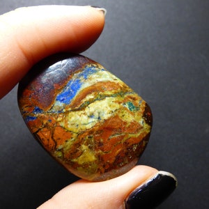 May include: A polished, oval-shaped stone with a mix of colors. The stone features bands of blue, brown, orange, and green. The stone is held between two fingers against a dark background. The stone is a natural mineral.