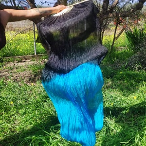 May include: A black and turquoise silk dance veil with a ruffled texture. The veil is held by a hand and is flowing in the wind.