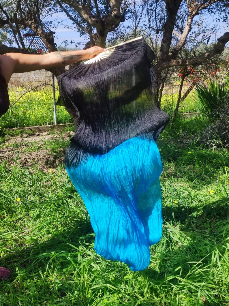 May include: A black and turquoise silk dance veil with a ruffled texture. The veil is held by a hand and is flowing in the wind.