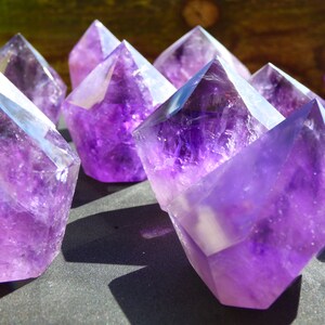 May include: A group of amethyst crystals, each with a pointed tip and a faceted, geometric shape. The crystals are a deep purple colour and have a translucent quality. They are arranged on a dark grey surface.