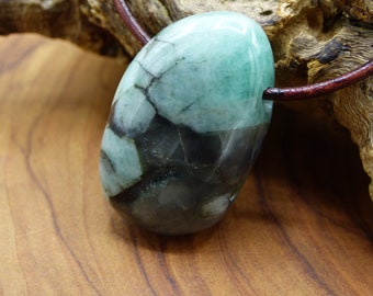 May include: A polished, oval-shaped emerald pendant featuring a mix of translucent light green and opaque dark grey to black inclusions, suspended on a thin, dark brown leather cord that passes through a hole drilled near the top edge.