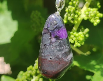 May include: A polished, irregular, teardrop-shaped pendant featuring a mix of deep purple, dark charcoal, and reddish-brown mineral inclusions. The stone is suspended from a simple silver-toned metal chain with a polished bail attachment.