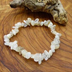 Può includere: Un braccialetto composto da piccole pietre chiare di forma irregolare. Le pietre sono infilate insieme per formare una forma circolare. Il braccialetto è appoggiato su una superficie di legno con un pezzo di legno alla deriva sullo sfondo.