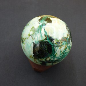 Pode incluir: Uma pedra decorativa esférica com uma mistura de cores branca, verde e castanho escuro. A pedra está sobre uma pequena base retangular castanha. O efeito geral é uma estética natural e terrosa.