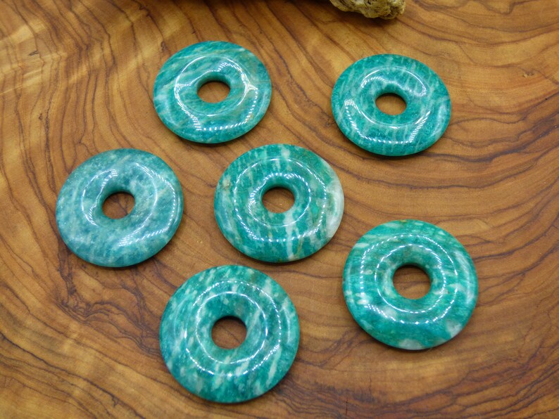 Pode incluir: Seis pedras de aventurina verde em forma de donut com um furo no centro. As pedras est&atilde;o dispostas em c&iacute;rculo sobre uma superf&iacute;cie de madeira.