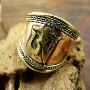 Puede incluir: Un anillo ancho de cobre con diseño y ribete de latón. El anillo presenta un emblema central de latón sobre una banda de cobre, enmarcado por un borde de latón trenzado. El anillo se exhibe sobre un trozo de madera.