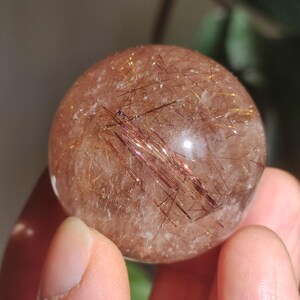 Könnte beinhalten: Eine klare Quarzkugel mit roten und goldenen Einschlüssen. Die Kugel ist etwa so groß wie ein Golfball und wird in einer Hand gehalten.