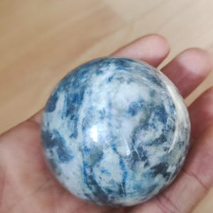 Puede incluir: Una esfera azul y blanca, sostenida en una mano. La esfera tiene un aspecto veteado con vetas azules y blancas. La superficie es lisa y reflectante, lo que sugiere una piedra o cristal pulido.