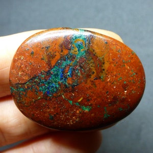 May include: A polished, oval-shaped stone with a rich, earthy color palette. The stone features a rusty-red base, speckled with small green and blue mineral deposits. The surface is smooth and reflective, showcasing natural patterns and textures.