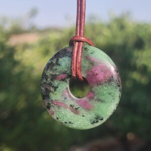 Può includere: Un ciondolo rotondo in zoisite lucidata con un foro centrale, sospeso a un cordino in pelle marrone. La pietra presenta un mix di colori verdi, rosa e neri, creando un motivo unico. Il ciondolo è una pietra preziosa naturale.