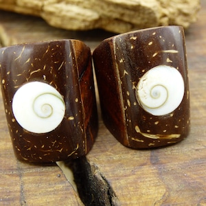 May include: Two dark brown, rectangular rings with a white spiral design in the center. The rings are made of a natural material, possibly wood or coconut shell, and sit on a wooden surface. The rings are likely jewelry.