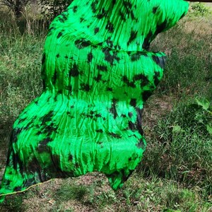 May include: Bright green fabric with black tie-dye patterns. The fabric is ruffled and flowing, with a yellow trim along the edge. The fabric is being held up in front of a grassy background.