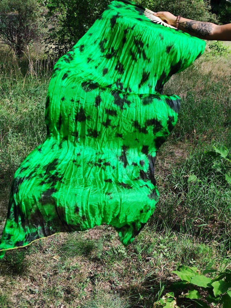 May include: Bright green fabric with black tie-dye patterns. The fabric is ruffled and flowing, with a yellow trim along the edge. The fabric is being held up in front of a grassy background.