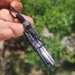 May include: A black kyanite pendant necklace. The kyanite crystal is a dark, metallic black with a textured surface. The pendant is suspended from a black cord with a decorative silver-colored bead at the top.