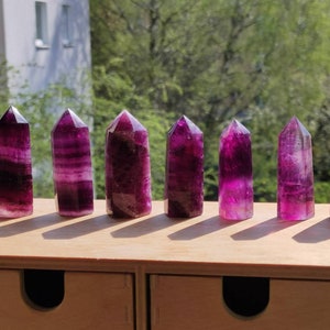 Può includere: Otto punte di cristallo di fluorite viola e bianche disposte in fila su una superficie di legno. I cristalli sono tutti di dimensioni e forma approssimativamente uguali e hanno una finitura liscia e lucida.