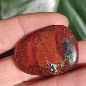May include: A red, green, and white jasper stone with a smooth, polished surface. The stone is oval-shaped and has a natural, organic pattern.