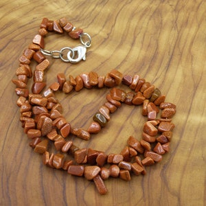 May include: A necklace made of small, irregular, reddish-brown stones with a metallic shimmer. The necklace is strung together with a silver-colored clasp. The stones are scattered across a wooden surface.